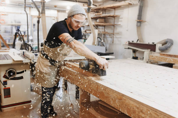 carpenter working with electric planer on wooden plank in workshop.